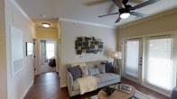 This single-floor residence centers around a welcoming living room designed for comfort and style. The space features warm, neutral-toned walls accented by crisp crown molding that adds a refined architectural touch. A ceiling fan with a built-in light fixture anchors the room, providing both function and visual focus overhead. Rich, dark hardwood flooring grounds the space and contrasts beautifully with the light walls, creating a warm and inviting ambiance.

The living area is furnished with a plush gray sofa adorned with decorative pillows and a soft yellow throw blanket, positioned in front of a round wooden coffee table. A striking abstract wall piece in dark and natural wood tones serves as a focal point, adding artistic character to the room. A tripod floor lamp in the corner provides warm ambient lighting, complementing the natural light that filters through a large window fitted with white horizontal blinds.

A hallway visible from the living room leads to a carpeted bedroom, hinting at a well-connected and functional floor plan. The overall ambiance is cozy, modern, and thoughtfully curated throughout.