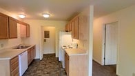 This single-floor space centers around a galley-style kitchen that runs along one wall, making efficient use of the available area. The kitchen is outfitted with light wood cabinetry both above and below the countertops, providing ample storage. White appliances, including an electric coil range and a full-size refrigerator, are integrated seamlessly into the layout. The countertops are light-colored and offer a clean, functional workspace. The flooring throughout the kitchen features a dark stone-tile pattern, adding texture and visual contrast to the neutral surroundings. Walls are painted in a soft, warm white that contributes to a bright and open feel. A doorway visible at the end of the kitchen leads to an adjacent utility or storage area, where an electrical panel is accessible. Additional doorways branch off from the kitchen, connecting to other areas of the space and suggesting a straightforward, open floor plan. Flush-mount ceiling lighting provides consistent illumination throughout. Overall, the space offers a clean, no-frills design focused on practicality and ease of use.