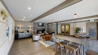 This single-floor space showcases a thoughtfully designed open-concept layout that seamlessly integrates a lounge and dining area into one cohesive environment. The room is anchored by a striking stacked-stone fireplace topped with a wall-mounted television, serving as a natural focal point. Reclaimed-style wood paneling lines one wall, adding warmth and a rustic character that complements the neutral color palette throughout.

The lounge area features plush seating arranged around the fireplace atop a patterned area rug, creating an inviting gathering space. Adjacent to it, a communal dining table with wooden barstools provides a more casual seating option, ideal for group use. The two zones flow naturally into one another without visual barriers, making the space feel open and versatile.

Exposed ceiling beams in a taupe finish add architectural interest overhead, while recessed lighting and flush-mount fixtures ensure the space is well-lit. A decorative metal sideboard near the lounge adds an industrial touch. A glass-paneled door in the background hints at an additional room beyond, suggesting further functional space. Light hardwood flooring ties the entire area together with a clean, contemporary feel.