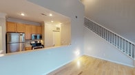 This two-story residence is defined by its dramatic double-height living room, where a soaring ceiling and a generous bank of stacked windows flood the space with natural light and views of surrounding greenery. A staircase with a classic white-railed banister anchors one side of the room, while light-toned hardwood flooring extends throughout the main level, lending warmth and continuity.

The kitchen is positioned off the living area in a semi-open layout, separated by a raised breakfast bar. It features light maple cabinetry with crown molding detail, stainless steel appliances including a refrigerator, microwave, and range, and ample counter space for everyday use. A front entry door is visible just beyond the kitchen, providing a practical flow from entry to living space.

Upstairs, the bedrooms are carpeted and comfortably sized, each featuring natural light from individual windows and dedicated closet space. One bedroom includes a walk-in closet outfitted with wire shelving, offering generous storage capacity.

Two full bathrooms serve the residence, each finished with tile flooring, a tub-shower combination, a single-sink vanity with light wood cabinetry, and clean white fixtures throughout. The overall aesthetic is neutral and well-maintained, creating a move-in ready environment.