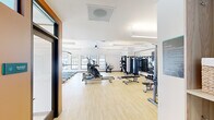 Located at 6609 E Warren Drive in Denver, CO, this fitness center offers a thoughtfully designed, single-floor space that provides a comprehensive workout environment. The open-plan layout is finished with light hardwood-style flooring throughout, contributing to a clean and energizing atmosphere.

The cardio zone, positioned along a wall of floor-to-ceiling windows, features a row of treadmills and elliptical machines from True Fitness, allowing users to exercise while taking in outdoor views and benefiting from ample natural light. A WaterRower rowing machine adds a premium touch to the cardio offerings.

The strength training area is equally well-appointed, with a full cable and functional trainer system, a multi-station cable machine, a complete dumbbell rack, and multiple adjustable benches. A large wall-mounted mirror with an integrated flat-screen TV enhances both the functionality and ambiance of this zone.

Overhead, modern ceiling fans and recessed linear lighting ensure comfortable climate control and consistent illumination throughout. The design incorporates warm terracotta accent walls near the entry vestibule, complemented by black-framed glass partitions, adding a contemporary architectural character to the space. The overall environment is bright, spacious, and purpose-built for an effective fitness experience.