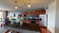 This single-floor space is thoughtfully designed to serve as a communal gathering area, blending a fully equipped kitchen with comfortable lounge and dining zones. The kitchen is outfitted with warm-toned wood cabinetry extending to the ceiling, dark granite countertops, stainless steel appliances including a refrigerator, range, and microwave, and a large island with pendant lighting overhead. Rich hardwood flooring anchors the kitchen area, transitioning to patterned carpet throughout the lounge and dining spaces.

The lounge area is defined by a wall-mounted flat-screen television, a freestanding pellet or wood stove, and mid-century modern-style seating including a sofa and accent chairs arranged around a round coffee table. Lush potted plants add warmth and life to the space. A standout feature is the expansive row of large windows along one wall, flooding the lounge with natural light and offering broad panoramic views of the surrounding cityscape.

The dining area accommodates multiple high-top tables with modern chairs, accented by decorative wall art including an ornate metal panel and an oversized clock. The overall color palette of gray, warm wood, and neutral tones creates a cohesive, contemporary ambiance suitable for socializing and relaxation.