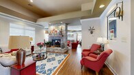 Located at 2945 Elam Rd in Murfreesboro, Tennessee, this single-floor community space is designed to foster comfort and social connection. The open layout flows seamlessly across multiple lounge areas, all unified by warm hardwood flooring and recessed lighting set into tray ceilings that add architectural depth and dimension throughout.

The centerpiece of the space is a striking floor-to-ceiling stone fireplace, which anchors the main seating area and creates a welcoming focal point. Surrounding it are plush red accent chairs, a patterned sectional sofa in neutral tones, and colorful area rugs that add vibrancy and definition to each zone. A bold blue-and-orange geometric rug anchors one of the primary lounge groupings, complemented by a glass-topped coffee table and coordinating side cabinets in deep red.

Toward one end of the space, a stone-faced bar counter with bar stools serves as a social hub, bathed in natural light from large windows fitted with white blinds and decorative valances. An additional seating area nearby features plaid furnishings and casual round tables, ideal for relaxed conversation.

Decorative touches—including wall-mounted artwork, a deer head accent, and carefully arranged lamps—lend the space a curated, lodge-inspired charm. Natural light floods in from multiple window banks, enhancing the warm, inviting ambiance of this thoughtfully designed communal retreat.