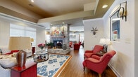 Located at 2945 Elam Rd in Murfreesboro, Tennessee, this single-floor community space is designed to foster comfort and social connection. The open layout flows seamlessly across multiple lounge areas, all unified by warm hardwood flooring and recessed lighting set into tray ceilings that add architectural depth and dimension throughout.

The centerpiece of the space is a striking floor-to-ceiling stone fireplace, which anchors the main seating area and creates a welcoming focal point. Surrounding it are plush red accent chairs, a patterned sectional sofa in neutral tones, and colorful area rugs that add vibrancy and definition to each zone. A bold blue-and-orange geometric rug anchors one of the primary lounge groupings, complemented by a glass-topped coffee table and coordinating side cabinets in deep red.

Toward one end of the space, a stone-faced bar counter with bar stools serves as a social hub, bathed in natural light from large windows fitted with white blinds and decorative valances. An additional seating area nearby features plaid furnishings and casual round tables, ideal for relaxed conversation.

Decorative touches—including wall-mounted artwork, a deer head accent, and carefully arranged lamps—lend the space a curated, lodge-inspired charm. Natural light floods in from multiple window banks, enhancing the warm, inviting ambiance of this thoughtfully designed communal retreat.