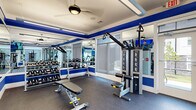 Located at 635 Bay Ave in Toms River, NJ, this single-story fitness center is thoughtfully designed to support a comprehensive workout experience. The space is defined by a striking blue-and-white color scheme, with bold royal blue accent bands lining the walls and contrasting cleanly against white shiplap paneling and mirrored surfaces that visually expand the room.

The fitness center is organized into distinct functional zones. The cardio area features a row of treadmills, elliptical trainers, and a stair climber, all positioned near large windows that invite natural light and provide views of the surrounding exterior. Wall-mounted flat-screen televisions are positioned throughout for entertainment during workouts.

The strength and free-weight area is equipped with a full dumbbell rack, kettlebells, adjustable benches, and cable resistance machines, including a multi-functional cable system suited for a variety of exercises. Additional functional training equipment rounds out the offerings.

The flooring throughout is durable rubber gym flooring, practical for both safety and equipment placement. Ceiling fans and fluorescent lighting ensure the space remains comfortable and well-lit. Glass-paned doors offer views into adjacent areas and contribute to an open, airy atmosphere across the entire facility.
