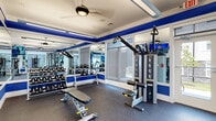 Located at 635 Bay Ave in Toms River, NJ, this single-story fitness center is thoughtfully designed to support a comprehensive workout experience. The space is defined by a striking blue-and-white color scheme, with bold royal blue accent bands lining the walls and contrasting cleanly against white shiplap paneling and mirrored surfaces that visually expand the room.

The fitness center is organized into distinct functional zones. The cardio area features a row of treadmills, elliptical trainers, and a stair climber, all positioned near large windows that invite natural light and provide views of the surrounding exterior. Wall-mounted flat-screen televisions are positioned throughout for entertainment during workouts.

The strength and free-weight area is equipped with a full dumbbell rack, kettlebells, adjustable benches, and cable resistance machines, including a multi-functional cable system suited for a variety of exercises. Additional functional training equipment rounds out the offerings.

The flooring throughout is durable rubber gym flooring, practical for both safety and equipment placement. Ceiling fans and fluorescent lighting ensure the space remains comfortable and well-lit. Glass-paned doors offer views into adjacent areas and contribute to an open, airy atmosphere across the entire facility.