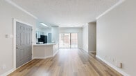 This single-floor, 2-bedroom, 2-bathroom residence offers a thoughtfully designed open-concept layout with modern finishes throughout. Light-toned wood-look flooring runs seamlessly through the main living areas, complemented by neutral warm-gray walls, crisp white baseboards, and textured popcorn ceilings with crown molding accents.

The combined kitchen and living room serves as the heart of the home. The kitchen features gray shaker-style cabinetry with brushed nickel hardware, stone-look countertops, and a curved breakfast bar peninsula. The open living area receives abundant natural light through a large sliding glass door fitted with vertical blinds, offering outdoor views and easy access.

The primary bedroom is a well-sized retreat with its own en-suite bathroom. This bathroom is equipped with a wide gray vanity, stone-look countertop, a lighted mirror, and wood-look flooring. The second bedroom is comfortable and naturally lit via a window with blinds. A second full bathroom includes a tub/shower combination with white tile surround and a matching gray vanity.

A central hallway connects all rooms efficiently, and the unit also includes three closets for ample storage. The overall ambiance is clean, bright, and move-in ready, reflecting a cohesive modern aesthetic throughout.