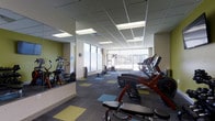 This fitness center offers a thoughtfully designed workout environment spread across a single floor. The space is anchored by a generous array of cardio equipment, including elliptical trainers, treadmills, stationary bikes, and rowing machines, all arranged to maximize usability and flow. A dedicated strength training area features a cable machine system, providing members with versatile resistance training options. Large floor-to-ceiling windows line one side of the space, flooding the interior with abundant natural light and offering views of the surrounding outdoor landscape — creating an open, motivating atmosphere. A full-length mirror wall on one side visually expands the space and supports functional movement and form checks. The ceiling is finished with a suspended tile grid fitted with recessed fluorescent lighting, maintaining consistent, even illumination throughout. The flooring features a multi-toned carpet tile pattern in green, blue, and neutral tones, adding a vibrant visual element to the space. Accent walls in a bold green tone complement the modern, clean aesthetic. The overall ambiance is bright, functional, and welcoming — well-suited for a range of fitness activities from light cardio to more intensive training sessions.
