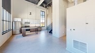 Located at 515 W Fourth St in Newton, Iowa, this two-floor industrial-style unit blends raw architectural character with contemporary updates. The main floor commands attention with soaring ceilings, exposed steel ductwork, and painted concrete floors that speak to the building's industrial heritage. Oversized, black-framed grid windows dominate the exterior walls, flooding the open living and kitchen area with natural light and offering views of the surrounding landscape. The kitchen is well-appointed with white cabinetry, stainless steel appliances including a range, microwave, and dishwasher, complemented by a sleek island with a dark wood base. A striking open-tread metal staircase with a black handrail connects the main floor to the upper level. Painted concrete block walls add texture and an authentic industrial aesthetic throughout. The upper floor offers a more intimate atmosphere with carpeted rooms, painted block walls, closet storage, and two full bathrooms finished with warm wood vanities, gray countertops, and large framed mirrors. The second floor also features additional rooms with large windows providing continued outdoor views. This space delivers a distinctive combination of open, loft-inspired living and functional private spaces.