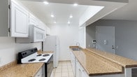 This single-floor residence offers a well-designed layout centered around a galley-style kitchen that maximizes efficiency and functionality. The kitchen is outfitted with white cabinetry that extends to the ceiling, providing generous storage space along both sides of the corridor. Granite-look laminate countertops run the length of the kitchen, offering ample workspace for meal preparation. White appliances, including a freestanding electric range, over-the-range microwave, and a full-size refrigerator, are neatly integrated into the cabinetry. A stainless steel sink with a modern faucet is positioned along the countertop, complemented by a window that brings in natural light. The kitchen flows openly toward an adjacent area, where a security panel and access door are visible, suggesting a thoughtful layout with direct entry access. Recessed lighting is installed throughout, including within a raised ceiling detail above the kitchen, creating a bright and well-lit environment. Light-toned ceramic tile flooring runs throughout, contributing to the clean, cohesive aesthetic of the space. Overall, the residence presents a practical and tidy interior with a neutral color palette and functional design details suited for comfortable everyday living.
