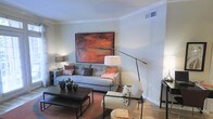 This single-floor space showcases a well-appointed living area designed with a cohesive modern aesthetic. The room is anchored by a neutral-toned sofa dressed with rust and patterned accent pillows, complemented by a matching upholstered armchair that adds an inviting seating arrangement. A bold abstract painting in warm earthy reds and browns serves as a striking focal point on the main wall, tying together the room's warm color palette.

Lighting is carefully considered, with a large arc floor lamp providing dramatic overhead illumination and a classic table lamp offering softer ambient light. Crown molding along the ceiling adds a refined architectural detail, elevating the overall finish of the space. The flooring transitions between carpet in the seating area and hard flooring near the perimeter, adding visual interest and defining zones.

A dark rectangular coffee table grounds the seating area, styled with small potted greenery and decorative objects for an organic, lived-in feel. The space balances clean, contemporary lines with warm tones and layered textures, resulting in an atmosphere that feels both polished and approachable.
