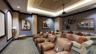 This dedicated home theater is a cinephile's dream, thoughtfully designed to deliver an immersive entertainment experience. The room features tiered seating arranged across multiple rows, offering unobstructed sightlines from every seat. Plush, oversized leather recliners and a large sectional sofa — accented with rich red and neutral throw pillows — provide a comfortable and stylish viewing environment.

The ceiling is a standout architectural element, showcasing an elegant tray design with ornate crown molding and integrated ambient LED lighting that casts a warm, atmospheric glow. A ceiling-mounted projector sits at the center, ready to deliver a large-format viewing experience on the front wall.

The décor pays clear homage to the golden age of Hollywood. Framed classic cinema portraits adorn the grey-toned walls, while oversized vintage film reels are mounted as dramatic decorative accents, reinforcing the cinematic theme throughout.

Soft, patterned carpeting in muted teal tones covers the floor, adding warmth and acoustic comfort to the space. Recessed lighting supplements the tray ceiling illumination, allowing for flexible mood control. The overall ambiance is sophisticated yet inviting, striking a perfect balance between a luxurious private lounge and a fully functional screening room.