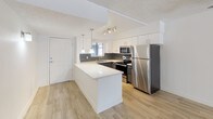 This single-floor space showcases a thoughtfully designed open-concept layout that seamlessly integrates living and kitchen areas. The kitchen is a standout feature, outfitted with sleek white upper and lower cabinetry that provides ample storage. A peninsula island with a crisp white quartz countertop extends the workspace and offers additional seating or prep space. The kitchen is equipped with a full suite of stainless steel appliances, including a refrigerator, electric range, and built-in microwave, complemented by a subway tile backsplash in a classic grey tone that adds texture and visual interest. Pendant lighting hangs above the peninsula, while a track lighting system illuminates the cooking area, ensuring excellent task lighting throughout. Light-toned laminate wood flooring runs throughout the space, enhancing the bright and airy feel. The textured white ceiling adds a subtle depth to the interior. Natural light filters in through a window above the sink, contributing to the overall sense of openness. The entry door is conveniently positioned near the kitchen, making for a practical and welcoming flow into the space. Overall, the design strikes a clean, contemporary aesthetic that is both functional and visually appealing.