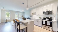 This thoughtfully designed single-floor residence offers a cohesive open-concept layout that maximizes its well-proportioned footprint. The kitchen is a standout feature, appointed with crisp white shaker cabinetry, white subway tile backsplash, stainless steel appliances, and a central island with bar seating — all illuminated by stylish globe pendant lights and a track lighting system overhead. Light hardwood-style flooring runs throughout, enhancing the bright, airy atmosphere.

The living room flows seamlessly from the kitchen and is bathed in natural light through large windows and a glass-paneled exterior door that frames views of a lush green landscape, creating a seamless indoor-outdoor connection. A ceiling fan adds practical comfort to the space.

The bedroom continues the home's light and neutral palette, featuring a generously sized window with the same scenic outdoor views, providing a calm and restful atmosphere. The bathroom is neatly finished with grey shaker-style cabinetry, a wide vanity with white countertops, and a large mirror.

A dedicated in-unit laundry room houses a full-size side-by-side washer and dryer, complemented by wire shelving and additional storage space. Every detail of this residence reflects a clean, modern aesthetic with functional design throughout.