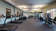 Located at 10315 Monrovia Dr in Matthews, NC, this fitness center is a well-equipped, single-floor facility designed to support a comprehensive workout experience. The space features rubber flooring throughout, providing both durability and comfort underfoot across all exercise zones.

The cardio area is generously outfitted with multiple treadmills and elliptical cross-trainers, each positioned to allow ample space between users. A wall-mounted flat-screen television is conveniently placed for entertainment during workouts. The strength training section includes a full power rack with weight plates, a dumbbell rack with a selection of free weights, and an adjustable bench, offering versatility for a wide range of resistance exercises. Additional cable and resistance machines round out the equipment offerings.

The facility is illuminated by a combination of pendant and linear overhead lighting, giving the space a bright, energetic atmosphere. Soft teal-toned walls add a calm, modern aesthetic throughout. Large mirrors along one wall enhance the sense of openness and allow users to monitor their form. A notable design feature is a large glass garage-style door that opens to the outdoors, inviting natural light and fresh air into the space.

Bold, large-scale artwork — including vivid portrait paintings — adds a dynamic and motivating visual character to the environment. Small personal lockers are available near the entrance for secure storage during visits.