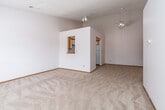 This single-floor space offers a practical and straightforward layout designed for comfortable everyday living. The main living area is generously sized, featuring neutral-toned carpet underfoot, white walls, and a textured ceiling. A notable architectural detail is the interior pass-through window built into a partial dividing wall, which visually connects the living area to the kitchen while maintaining a sense of defined spaces. Track lighting and a ceiling fan provide functional illumination throughout.

The kitchen features white cabinetry with warm wood-toned trim, white laminate countertops, a double-basin stainless steel sink, and a dishwasher. Tile flooring completes the kitchen's clean, utilitarian design. A white range is also present, providing full cooking functionality.

The bedroom areas are carpeted and bright, with at least one room benefiting from a large window that allows natural light and outdoor views. Additional rooms offer quiet, neutral spaces suitable for various uses.

The bathroom includes a tiled floor in warm earth tones, a full tub-and-shower combination with a safety grab bar, a vanity with counter space, and a toilet. Warm wood-toned doors throughout the unit add a consistent design element, contributing to an overall cohesive and functional aesthetic.