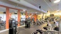 This fitness center offers a comprehensive workout environment designed to accommodate a variety of exercise routines. The space features an open floor plan with a generous selection of cardio equipment, including treadmills, elliptical trainers, and stationary bikes, all arranged to maximize accessibility and flow. Strength training stations and additional exercise machines are thoughtfully distributed throughout the room, providing options for both targeted and full-body workouts.

The interior design is characterized by bold orange accent columns and yellow walls, creating an energizing and motivating atmosphere. Exposed ductwork along the ceiling contributes an industrial aesthetic, while pendant lighting and track-mounted fixtures ensure the space is well-lit throughout. Large mirrors line the walls, visually expanding the room and allowing members to monitor their form during exercise.

Rubber flooring covers the workout area, offering both durability and comfort underfoot. Windows along one wall allow for natural light to filter in, enhancing the overall ambiance. The layout is practical and efficient, with clear sightlines across the floor, making it easy to navigate between equipment. Overall, this fitness center is a functional, well-appointed space suited for a broad range of fitness goals and skill levels.
