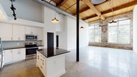 Located at 701 Easley Bridge Rd in Greenville, SC, this single-floor unit is a thoughtfully converted industrial space that seamlessly blends historic character with modern finishes. The architecture retains original elements including exposed brick walls, warm wood beam ceilings, visible ductwork, and structural steel columns, all complemented by polished concrete floors throughout.

The open-concept living area benefits from soaring ceilings and oversized factory-style windows framed by original brick, allowing generous natural light to permeate the space and offering views of the surrounding landscape. Ceiling fans and pendant lighting add functional ambiance to the expansive main area.

The kitchen is well-appointed with white cabinetry, dark countertops, stainless steel appliances, a tile backsplash, and a central island with additional storage—all beneath dramatic pendant lighting and elevated ceilings.

Two bathrooms provide modern comfort: one features a glass-enclosed walk-in shower, soaking tub, dark-hardware fixtures, and a vanity with dark countertops; the second offers a tiled tub-shower combination with matching black fixtures and a mirrored vanity. The overall aesthetic is a refined industrial style with a clean, contemporary sensibility.