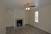 This single-story space presents a straightforward, functional layout with a clean and neutral aesthetic throughout. The main living area serves as the focal point of the home, anchored by a fireplace with a tile surround and raised tile hearth that adds a modest architectural detail to the otherwise understated room. The fireplace is framed by a smooth, white-painted chimney breast that extends to the ceiling, providing a natural visual centerpiece.

The flooring throughout the living area consists of wood-look planks in a warm, medium-toned finish, contributing warmth and continuity to the space. Light gray walls and white baseboards maintain a clean, cohesive look. A ceiling fan is partially visible in the upper corner, indicating practical climate control for the living area.

A hallway visible from the main room leads to the entry door, suggesting a transitional space between the living area and the entrance. The entry corridor features the same wood-look flooring, maintaining design consistency. A wall-mounted HVAC vent is visible near the ceiling, and the overall layout appears well-maintained and move-in ready, suited for comfortable everyday living.