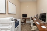 Located at 700 SR-96 E in Bonaire, Georgia, this single-floor business center offers a well-appointed workspace designed for efficiency and professional use. The space is finished with warm beige walls, crisp white trim, and patterned carpet tiles that lend a polished yet approachable atmosphere throughout.

The main workstation area features an L-shaped and straight-run configuration of rich wood-toned desks lined with multiple iMac computers and ergonomic rolling chairs, providing ample seating for several users simultaneously. Recessed lighting illuminates the workspace evenly, ensuring a comfortable environment for extended use.

A dedicated printing and equipment alcove houses commercial-grade multifunction printers and copiers, making the space fully equipped for document handling needs. This area is accented by a pair of framed textured wall art pieces, adding a refined aesthetic touch to an otherwise utilitarian corner.

Double panel doors with a clean white finish connect the business center to adjacent areas, enhancing accessibility and flow. The overall design balances functionality with a tidy, professional ambiance, making this business center a practical and well-resourced facility for residents or guests seeking a reliable workspace.