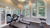 Located at 2422 Banks Edge Way in Reynoldsburg, Ohio, this single-floor fitness facility offers a thoughtfully designed space that combines functionality with refined architectural detail. The standout feature of the workout room is its series of large arched windows, which flood the interior with abundant natural light while framing picturesque views of the surrounding mature trees and lush greenery. The windows are elegantly trimmed in white, complementing the soft gray walls and creating a bright, airy atmosphere that makes exercising feel less like a chore and more like a retreat. The room is finished with durable rubber flooring, ideal for fitness use, and is equipped with treadmills positioned to take full advantage of the scenic outdoor views. Crown molding and a tray ceiling with recessed lighting add a polished, upscale character to the space, elevating it beyond a standard gym environment. A large mirror along one wall further enhances the sense of openness and allows for form-checking during workouts. The overall ambiance is clean, well-lit, and inviting, making this a premium fitness space that balances practical athletic use with an aesthetically pleasing, nature-connected setting.