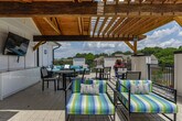 Located along Atlanta Student Movement Boulevard SW in Atlanta, Georgia, this rooftop terrace offers an inviting outdoor retreat designed for both relaxation and social gatherings. The space is thoughtfully divided into distinct seating areas, each serving a unique purpose while maintaining a cohesive aesthetic throughout.

A partially covered lounge area anchors one side of the terrace, featuring exposed wood beam ceiling accents that add warmth and architectural character. This shaded zone is furnished with a generously sized sectional sofa in teal upholstery, complemented by colorful striped accent chairs and a central fire pit table — creating an ideal setting for casual conversation and evening gatherings.

Adjacent to the lounge, an open-air dining area provides a round table with seating for multiple guests, bathed in natural sunlight and framed by vibrant potted flowers that add a lively pop of color. Light-toned tile flooring extends across the entire terrace, reflecting sunlight and contributing to the bright, airy atmosphere.

The terrace is enclosed by sleek black metal railings, offering unobstructed panoramic views of the Atlanta skyline, lush tree canopies, and the surrounding residential community. Built-in cabinetry with a stone countertop provides additional functionality along one wall. The combination of open sky, greenery, and thoughtful furnishings makes this rooftop terrace a standout feature of the property.