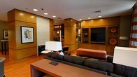 This single-floor space presents a thoughtfully designed lounge or common area with a refined, contemporary aesthetic. The interior is defined by warm honey-toned wood paneling that lines the walls and structural columns, lending a sense of cohesion and sophistication throughout. Recessed lighting embedded in the white ceiling illuminates the space evenly, complementing the overall polished ambiance.

The seating area is anchored by a dark charcoal sofa paired with a bold orange upholstered ottoman, creating a striking contrast against the warm wood tones. A large floor lamp with a rectangular white shade adds ambient lighting near the conversation area, while a low wooden coffee table grounds the arrangement.

A built-in entertainment wall dominates one side of the room, housing a flat-screen television alongside display shelving with glass-fronted cabinetry — ideal for storing books, décor, or media accessories. Further into the space, a secondary seating nook with a small round table and dark leather chair offers a more intimate setting. Decorative shelving units and subtle artwork add visual interest without overwhelming the clean, structured design. The hardwood flooring ties each zone together seamlessly.