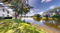 Located at Lakepointe Landing in Plantation, Florida, this space offers a remarkable connection to its natural surroundings. The standout feature is the sweeping waterfront view, where a tranquil lake stretches out and reflects the bright Florida sky, creating a calm and picturesque atmosphere. Mature trees frame the shoreline, their canopies providing natural shade and adding to the lush, tropical character of the setting. The grounds are carpeted with vibrant green grass that leads gently down to the water's edge, offering an inviting outdoor environment. Across the lake, residential structures are visible nestled among additional mature trees, reinforcing the peaceful, community-oriented character of the Lakepointe Landing neighborhood. The overall ambiance is defined by natural light, open skies dotted with clouds, and the serene quality of still water. The location in Plantation, FL situates this space within a well-established South Florida community known for its greenery and lakeside character. Whether enjoyed as an outdoor area or as a vantage point for the surrounding landscape, this space delivers an exceptional waterfront experience defined by natural beauty and tranquility.