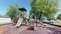 Located at Coventry Village Green Court in Newark, Ohio, this outdoor recreational space serves as a community playground within a well-established neighborhood setting. The centerpiece of the space is a multi-level play structure finished in green and tan, designed to accommodate children of various ages. The structure features multiple access points, including a traditional staircase and a rock-climbing wall, leading to an elevated platform topped with a covered canopy. From the platform, children can enjoy a curved slide as one of several descent options.

Surrounding the base of the play structure is a rubber mulch surface, providing a safety-conscious ground cover beneath the equipment. Adjacent to the main structure, a set of ground-level interactive elements — including mushroom-shaped stepping stools — offers additional play opportunities for younger children.

The playground is framed by a generous canopy of mature deciduous trees, which provide natural shade and contribute to a welcoming, park-like atmosphere. The open sky and lush greenery visible throughout the space create a bright, inviting outdoor environment. Nearby parking is accessible, making the space convenient for families throughout the Coventry Village Green community.