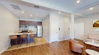 Located at 1800 Stokes St in San Jose, California, this single-floor space combines a fully equipped kitchen and an open living area into a cohesive, efficiently designed layout. The kitchen is outfitted with rich dark mahogany cabinetry, stainless steel appliances including a refrigerator, electric range, and over-the-range microwave, complemented by a striking mosaic tile backsplash and light quartz countertops. A breakfast bar with seating provides a casual dining option, seamlessly connecting the kitchen to the living area. The living room features warm hardwood-style flooring, recessed lighting, and exposed ceiling beams that add architectural character. A set of full-length glass French doors floods the space with natural light and offers direct views of an outdoor pool area, creating an inviting indoor-outdoor connection. A wall-mounted flat-screen television, cozy leather accent chairs, and a neutral sofa arrangement make the living area both functional and comfortable. A structural column adds a subtle design element while an abstract wall painting introduces a touch of artistic flair. The overall ambiance is modern, clean, and welcoming, making this space well-suited for both everyday living and casual entertaining.