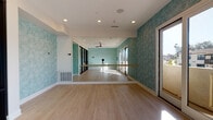 Located on Telegraph Road in Ventura, California, this single-floor yoga studio offers a thoughtfully designed environment built to support movement, mindfulness, and wellness. The main practice room is expansive and open, featuring smooth hardwood flooring that provides both warmth and functionality for yoga and barre classes. A full-length mirrored wall runs along one side of the studio, accompanied by a mounted wooden ballet barre — ideal for barre-style training and alignment practice. The walls are adorned with a distinctive teal textured wallcovering that lends a calming, serene atmosphere to the space. Recessed lighting and a ceiling fan ensure the room remains comfortable and well-lit during classes. Natural light streams in through large windows and a striking floor-to-ceiling black-framed glass partition wall, which visually connects the practice room to the adjacent lounge and reception area. The lounge features light large-format tile flooring, soft teal-painted walls, modern seating, and a sculptural amber pendant light overhead. A wall-mounted flat-screen display and decorative metal wall art complete the inviting, contemporary aesthetic. The glass partition allows for an open, transparent feel between the functional zones of the studio, creating a cohesive and welcoming space from entry to practice floor.