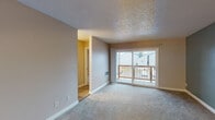 This single-floor space is thoughtfully laid out to maximize functionality across its key living areas. The main living room is finished with neutral-toned carpet and features a textured ceiling, white baseboards, and a sliding glass door that opens onto a wooden balcony, offering outdoor views and natural light. A wall-mounted heating unit provides year-round comfort in this central space.

Adjacent to the living area, the kitchen is finished with light tile flooring, warm oak cabinetry, speckled granite countertops, and white appliances including an electric range and built-in microwave. The transition between the kitchen and living spaces is defined by a change in flooring material, adding visual distinction.

The bedroom features soft carpet underfoot, neutral walls, and a textured ceiling consistent with the rest of the unit. A closet door provides practical storage within the room. An additional room, painted in a muted sage green, offers versatility and is similarly carpeted with hardwood-style flooring accenting the entryway.

The bathroom includes a clean, white tub and shower surround with built-in shelving and a brushed nickel towel bar, contributing to a simple, functional finish throughout the space.