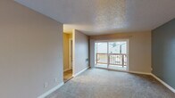This single-floor space is thoughtfully laid out to maximize functionality across its key living areas. The main living room is finished with neutral-toned carpet and features a textured ceiling, white baseboards, and a sliding glass door that opens onto a wooden balcony, offering outdoor views and natural light. A wall-mounted heating unit provides year-round comfort in this central space.

Adjacent to the living area, the kitchen is finished with light tile flooring, warm oak cabinetry, speckled granite countertops, and white appliances including an electric range and built-in microwave. The transition between the kitchen and living spaces is defined by a change in flooring material, adding visual distinction.

The bedroom features soft carpet underfoot, neutral walls, and a textured ceiling consistent with the rest of the unit. A closet door provides practical storage within the room. An additional room, painted in a muted sage green, offers versatility and is similarly carpeted with hardwood-style flooring accenting the entryway.

The bathroom includes a clean, white tub and shower surround with built-in shelving and a brushed nickel towel bar, contributing to a simple, functional finish throughout the space.