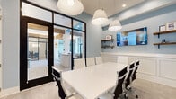 This contemporary conference room offers a well-appointed setting designed for productive meetings and collaboration. The space is defined by its striking floor-to-ceiling black-framed glass walls, which provide a seamless visual connection to the adjacent common areas while maintaining a sense of enclosure and focus. Natural light filters through the glass partitions, brightening the interior and creating an open, airy atmosphere.

The room centers around a sleek, white rectangular conference table surrounded by ergonomic high-back chairs in white and black, comfortably accommodating a full team. Overhead, a series of large white pendant lights hang at varying heights, casting warm, even illumination across the workspace.

One wall features a wall-mounted flat-screen display flanked by rustic wooden shelves adorned with decorative accents, adding warmth and character to the otherwise clean, modern aesthetic. An additional wall showcases a pair of framed abstract artwork pieces in coordinating blue tones, complementing the soft blue-gray paint that envelops the room.

Built-in white wainscoting runs along the lower portion of the walls, lending a refined, polished finish. Light wood-tone flooring ties the design together, balancing the crispness of the white furnishings with a natural, grounded feel. A direct exterior door provides convenient independent access, making this conference room both functional and sophisticated.