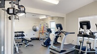 This fitness center at 5325 N Oak St in Springdale, Arkansas, is a compact yet thoughtfully outfitted exercise facility designed to support a wide variety of workout routines. The single-room space features durable rubber flooring throughout, providing both safety and comfort underfoot. Bright fluorescent lighting illuminates the entire area evenly, while multiple windows fitted with blinds allow natural light to enter and contribute to an open, energizing atmosphere.

The cardio section is anchored by TRUE-brand treadmills and an elliptical trainer, complemented by a stationary upright bike and a rowing machine, offering diverse options for aerobic exercise. A multi-station cable machine serves as the centerpiece of the strength training area, alongside a adjustable weight bench and a full rack of TRUE-branded dumbbells in graduated sizes. A wall-mounted flat-screen television adds an entertainment element for users during workouts.

The functional training corner is well-stocked with a stability ball, medicine balls of varying weights arranged on a dedicated rack, resistance bands, and a foam roller — catering to stretching, balance, and rehabilitation needs. A large wall mirror enhances the sense of space and allows users to monitor their form. The room is accessed via an exterior door and is equipped with a clearly marked emergency exit, reflecting attention to safety standards.
