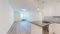 This well-designed two-bedroom, two-bathroom residence offers a clean, contemporary aesthetic across its thoughtfully laid-out single floor. Light hardwood-style flooring runs throughout the main living areas and bedrooms, creating a cohesive and bright atmosphere. The open-concept kitchen flows seamlessly into the living room, making the space feel open and connected. The kitchen is well-appointed with sleek white cabinetry, a gray tile backsplash, dark granite countertops, and a full suite of stainless steel appliances including a range, microwave, dishwasher, and a large side-by-side refrigerator. A peninsula with pendant lighting above serves as a natural divider between the kitchen and living area. The living room receives natural light through a window and provides a generous, open area. Both bedrooms are comfortably sized, each featuring windows with natural light and individual climate control units. The primary bedroom offers a notably spacious layout. The two bathrooms feature a refined design with marble-look tile flooring, a floating vanity with a white countertop, and a glass-enclosed walk-in shower with full-height tile surround. A hallway connects the rooms, providing a practical and organized floor plan throughout.