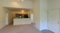 This single-floor space features an open-concept design that seamlessly connects the living area to the kitchen. The living area is finished with neutral wall-to-wall carpeting, contributing to a warm and comfortable atmosphere. Notably high ceilings enhance the sense of openness and volume throughout the main living space, allowing light to spread generously across the room.

The kitchen is positioned on a raised platform and separated from the living area by a breakfast bar-style counter finished with a dark countertop, creating a natural boundary while maintaining visual connectivity. The kitchen is equipped with warm-toned wooden cabinetry, dark countertops, and black appliances, including a microwave and range, offering a cohesive and practical cooking space.

A hallway visible beyond the kitchen leads to additional areas of the unit, suggesting a well-organized floor plan. A white paneled door on the right side of the living area provides access to a separate room or closet. A floor vent indicates a central HVAC system. The overall color palette of cream walls, neutral carpeting, and wood-toned cabinetry creates a cohesive and inviting environment throughout the space.