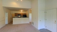 This single-floor space features an open-concept design that seamlessly connects the living area to the kitchen. The living area is finished with neutral wall-to-wall carpeting, contributing to a warm and comfortable atmosphere. Notably high ceilings enhance the sense of openness and volume throughout the main living space, allowing light to spread generously across the room.

The kitchen is positioned on a raised platform and separated from the living area by a breakfast bar-style counter finished with a dark countertop, creating a natural boundary while maintaining visual connectivity. The kitchen is equipped with warm-toned wooden cabinetry, dark countertops, and black appliances, including a microwave and range, offering a cohesive and practical cooking space.

A hallway visible beyond the kitchen leads to additional areas of the unit, suggesting a well-organized floor plan. A white paneled door on the right side of the living area provides access to a separate room or closet. A floor vent indicates a central HVAC system. The overall color palette of cream walls, neutral carpeting, and wood-toned cabinetry creates a cohesive and inviting environment throughout the space.
