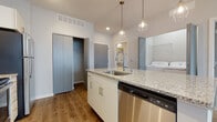 This well-appointed single-floor unit offers a thoughtfully designed open-concept layout that seamlessly blends the kitchen and living areas. The kitchen serves as the centerpiece of the space, anchored by a large island with a granite countertop, undermount sink, and stainless steel dishwasher. Geometric pendant lights hang above the island, adding a modern touch. The kitchen is further equipped with white shaker-style cabinetry, granite countertops along the perimeter, and a full suite of stainless steel appliances including a refrigerator, electric range, and built-in microwave. Track lighting illuminates the cooking area, while warm hardwood-style flooring flows throughout the main living spaces. A dedicated laundry area with a washer and dryer is conveniently accessible from the kitchen. The bedroom is a quiet, carpeted retreat featuring a ceiling fan, a generous window with blinds that welcomes natural light and outdoor views, and a spacious walk-in closet outfitted with wire shelving for organized storage. The bathroom features granite countertops on the vanity, a single undermount sink, a large mirror, and a walk-in shower with a clean white surround. The unit's neutral color palette and modern finishes create a bright, cohesive, and move-in-ready atmosphere throughout.