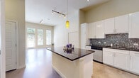 This single-floor space showcases a contemporary design highlighted by soaring ceilings and polished concrete floors that run throughout, creating a sleek, cohesive aesthetic. The open-concept layout connects the kitchen and living areas seamlessly, making the space feel expansive and airy.

The kitchen is well-appointed with crisp white cabinetry extending to the ceiling, dark granite countertops, and a striking mosaic tile backsplash featuring metallic tones. A central island with a matching dark countertop provides additional prep and gathering space. Stainless steel appliances, including a range, microwave, refrigerator, and dishwasher, complete the functional and stylish kitchen.

The living area benefits from tall ceilings and large windows alongside a glass-paneled exterior door, allowing generous natural light and outdoor views. Track lighting offers additional illumination throughout the main living space.

The bedroom is a calm, bright retreat with large windows that frame outdoor greenery and polished concrete floors continuing the home's consistent design language.

The bathroom features a dual-sink vanity topped with marble-patterned countertops, white cabinetry, a large mirror, and access to a walk-in closet, combining practicality with refined finishes throughout.