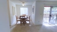 This single-floor space features a well-lit dining area that serves as a welcoming gathering spot. The room is finished with plush wall-to-wall carpeting in a neutral beige tone, complementing the clean white walls throughout. A round dark-wood dining table is centrally positioned, surrounded by four cushioned chairs with cross-back detailing, offering a classic and functional seating arrangement. Natural light filters into the space through a large window dressed with vertical blinds, casting warm sunlight across the floor and creating an airy atmosphere. A ceiling fan with an integrated light fixture is mounted overhead, providing both ventilation and ambient lighting. Framed artwork adorns the walls, adding a touch of character to the otherwise understated décor. A half-wall partition defines the dining area from an adjoining space, contributing to a sense of architectural structure while maintaining an open feel. A sliding or secondary access point visible to the right hints at an outdoor area, enhancing the overall connectivity of the interior to the exterior. The space presents a clean, functional, and comfortable environment.