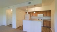 This single-floor space features an open-concept kitchen that serves as a central hub of the layout. The kitchen is equipped with warm-toned wooden upper cabinetry that provides ample storage, complemented by a suite of white appliances including a refrigerator, electric range, and built-in microwave. A peninsula with a light-toned countertop extends into the living area, creating a natural divide between the kitchen and adjacent spaces while offering additional workspace and seating potential. Two pendant lights hang above the peninsula, adding a practical yet stylish focal point to the area. The flooring in the kitchen transitions from tile to carpet as it moves into the surrounding living areas, delineating functional zones throughout the space. Neutral wall tones keep the interior feeling bright and clean, while a ceiling vent ensures comfortable climate control. A paneled interior door is visible from the kitchen, hinting at additional rooms beyond. Overall, the space delivers a straightforward and functional design with a cohesive, understated aesthetic.