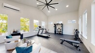 Located at 1471 SE Blueberry Rd in Port Orchard, WA, this fitness and lounge facility offers a bright, modern environment designed for both exercise and relaxation. The space is defined by soaring vaulted ceilings that create an open, airy atmosphere, complemented by a large statement ceiling fan with recessed lighting throughout. Expansive windows on multiple walls flood the interior with natural light and provide views of the surrounding greenery and neighborhood.

The fitness area features durable dark flooring and is outfitted with a treadmill, stationary cycle, multi-function cable machine, an adjustable weight bench, and a full rack of dumbbells — providing a comprehensive workout experience. A wall-mounted mini-split system ensures year-round climate comfort during exercise sessions.

Seamlessly integrated within the same space is a welcoming lounge area with light hardwood-style flooring, plush white accent chairs accented with teal throw pillows, and a round wooden coffee table. Adjacent to the lounge is a kitchenette with warm wood cabinetry, countertop space, and small appliances, making the space equally suitable for socializing or unwinding. A glass-panel entry door connects the interior to the outdoors, enhancing the sense of openness throughout.