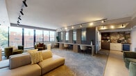 This thoughtfully designed single-floor amenity space blends comfort and functionality in a sleek, contemporary setting. The open layout is anchored by a generously sized lounge area furnished with a low-profile modular sofa, plush accent chairs in deep olive velvet, and side tables that create an inviting atmosphere for relaxation or casual gatherings. A wall of large floor-to-ceiling windows runs along the perimeter, flooding the space with natural light and offering views of the surrounding urban streetscape and greenery beyond.

Along one wall, a built-in co-working station features a continuous wood-toned desk surface paired with modern white chairs, divided by slatted wood paneling and framed mirrors that add depth and visual interest. Track lighting runs throughout the ceiling, providing layered and adjustable illumination across all zones.

Interior glass windows connect to an adjacent fitness area, maintaining visual openness while defining separate functional spaces. A corridor leads to a kitchenette and additional rooms, with built-in shelving and rich stone-accented wall details adding warmth and texture. The flooring transitions between soft carpet in the lounge areas and smooth hard surface in the corridor, reinforcing the distinction between zones. Overall, the space strikes a refined balance between a professional work environment and a comfortable residential retreat.