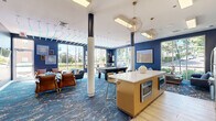 Located at 28 Southfield Ave in Stamford, Connecticut, this single-floor community lounge space is designed with a modern, upscale aesthetic that prioritizes both comfort and social engagement. The interior is defined by a striking navy blue color palette across the walls, complemented by crisp white ceilings and warm accent lighting from geometric gold pendant fixtures that hang elegantly overhead. The ceiling features an eye-catching linear light installation with blue-toned accents, adding a contemporary edge to the space. Floor-to-ceiling windows line multiple walls, flooding the interior with natural light and offering open views of the surrounding neighborhood. Structural columns wrapped in textured rope detailing serve as distinctive architectural elements throughout the room. The lounge area is furnished with plush leather sofas and armchairs in rich cognac and navy tones, arranged around coffee tables to create inviting conversation nooks. A full-size pool table anchors the entertainment zone, while a central island with a white quartz countertop and built-in appliances provides a functional kitchenette area with bar seating. A vibrant blue abstract-patterned carpet ties the entire space together, enhancing its dynamic, resort-style ambiance. Curated wall art and decorative accessories complete the polished, welcoming atmosphere.