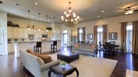 This single-story home showcases a beautifully designed open-concept floor plan that seamlessly connects the main living areas. The expansive living room serves as the heart of the home, anchored by a striking wrought-iron chandelier with frosted glass shades that casts warm light throughout the space. Recessed lighting complements the chandelier, illuminating the tall ceilings that lend an airy, grand feel to the interior. Rich dark hardwood floors run throughout, providing an elegant contrast to the neutral, light-toned walls and furnishings. The living area is generously sized, accommodating multiple seating arrangements centered around a tufted leather ottoman. Large windows dressed with floor-length draping curtains allow abundant natural light to fill the space while adding a refined decorative touch. The kitchen flows naturally from the living area and features white cabinetry, stainless steel appliances, and a breakfast bar with tall seating — ideal for casual dining. A dedicated dining area is visible beyond, offering additional space for formal meals. The overall ambiance is warm, sophisticated, and welcoming, blending classic design elements with a comfortable, livable layout throughout the single level.