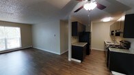 This single-floor space offers a practical and open layout that seamlessly connects the living area to the kitchen, creating an efficient and cohesive environment. The flooring throughout is a rich, dark hardwood-style material that adds warmth and a modern touch to the interior.

The living area is spacious and open, featuring neutral-toned walls that provide a clean, versatile backdrop. A ceiling fan with a built-in light fixture is centrally mounted, offering both airflow and ambient lighting. A baseboard heater is also present along the wall, ensuring comfort year-round.

The kitchen is defined by dark cabinetry with a bold, contemporary aesthetic. Upper and lower cabinets provide ample storage, while the light-toned countertop with subtle veining creates an appealing contrast against the darker cabinetry. A full-size black refrigerator completes the kitchen's cohesive color scheme. The kitchen is partially separated from the living area by a partial wall, maintaining a sense of openness while still defining the space.

The textured popcorn ceiling is consistent throughout, and recessed lighting adds supplemental illumination. Overall, this space presents a functional and straightforward layout with a modern, updated interior aesthetic suitable for everyday living.