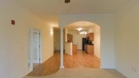 This single-floor residence showcases a thoughtfully designed open layout that seamlessly connects the living and kitchen areas. A defining architectural feature is the elegant arched entryway, framed by a decorative column, which creates a natural transition between spaces while maintaining an open, airy feel throughout.

The kitchen is well-appointed with warm honey-toned cabinetry, a peninsula that provides additional counter space and casual seating potential, and black appliances including a refrigerator and range. A pendant chandelier with multiple lights serves as a stylish focal point above this area, adding a touch of elegance to the functional space.

The flooring transitions from hardwood-style planks in the kitchen and main living areas to plush carpet in the living room, offering both practicality and comfort underfoot. Neutral cream-toned walls throughout create a bright, welcoming atmosphere. A ceiling fan is also present, contributing to year-round comfort. A doorway with glass-panel detail is visible to the left, hinting at additional rooms and further enhancing the overall sense of space and natural light within the home.