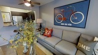 This two-floor residence offers a thoughtfully designed layout that blends comfort and style across its living spaces. The main living area serves as the heart of the home, featuring warm hardwood-style flooring, a ceiling fan overhead, and soft, neutral wall tones that create a welcoming atmosphere. A standout decorative piece — a bold, vintage-style "faire du vélo" bicycle artwork in rich blues and reds — anchors the living room wall, adding a pop of character and personality to the space. The living area is furnished with a plush, light gray sofa accented with vibrant throw pillows, complemented by a classic table lamp that adds warm ambient lighting. A vase of yellow florals on the dining surface introduces a natural, lively touch. The open-concept design allows for a seamless transition between the living and dining areas. Visible through an archway, the kitchen showcases dark espresso cabinetry, stainless steel appliances, and ample counter space, offering both functionality and a sleek aesthetic. The cohesive design language across the main floor balances modern finishes with inviting, homey details, making this a well-appointed and versatile living space spread across two floors.