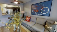 This two-floor residence offers a thoughtfully designed layout that blends comfort and style across its living spaces. The main living area serves as the heart of the home, featuring warm hardwood-style flooring, a ceiling fan overhead, and soft, neutral wall tones that create a welcoming atmosphere. A standout decorative piece — a bold, vintage-style "faire du vélo" bicycle artwork in rich blues and reds — anchors the living room wall, adding a pop of character and personality to the space. The living area is furnished with a plush, light gray sofa accented with vibrant throw pillows, complemented by a classic table lamp that adds warm ambient lighting. A vase of yellow florals on the dining surface introduces a natural, lively touch. The open-concept design allows for a seamless transition between the living and dining areas. Visible through an archway, the kitchen showcases dark espresso cabinetry, stainless steel appliances, and ample counter space, offering both functionality and a sleek aesthetic. The cohesive design language across the main floor balances modern finishes with inviting, homey details, making this a well-appointed and versatile living space spread across two floors.